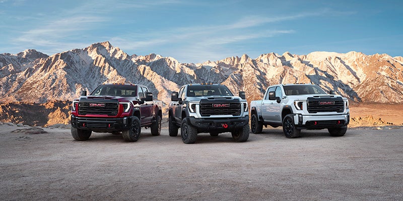 A red, silver, and white 2024 GMC Sierra 2500 HD parked alongside one another while the sun sets over mountains in the background